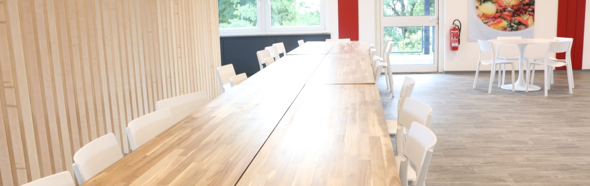 Blick auf den langen Holztisch der Cantina Rhein-Maas, links daneben der Raumtrenner aus Holzstreben und rechts daneben ein weißer Tisch mit fünf weißen Stühlen