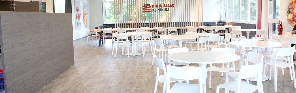 Blick in die Cantina des Rhein-Maas Klinikum mit sieben weißen Tischen und je fünf weißen Stühlen, einem langen Holztisch mit zwanzig Stühlen, Raumtrenner und großen Glasbildern an der der Wand