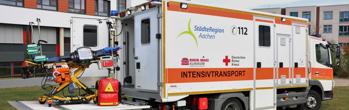 Ein Intensivtransportwagen steht vor dem Rhein-Massklinikum. Die hintere Tür steht offen und eine Patiententrage mit Überwachungsgeräten und ein roter Notfallrucksack stehen hinter dem Krankenwagen.