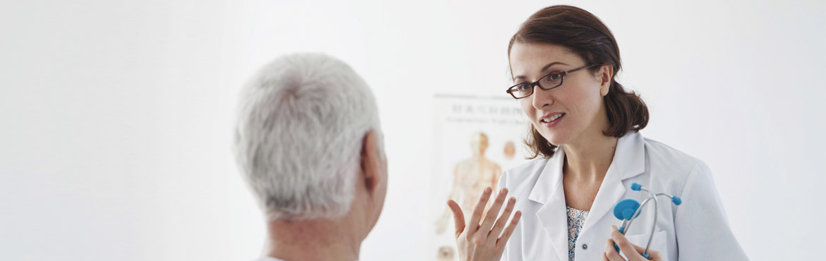 Eine jüngere Ärztin steht vor einem älterem Patienten und erklärt ihm etwas, in ihrer linken Hand hällt sie ein Stethoskop und hinter ihr an der Wand hängt ein anatomisches Plakat.