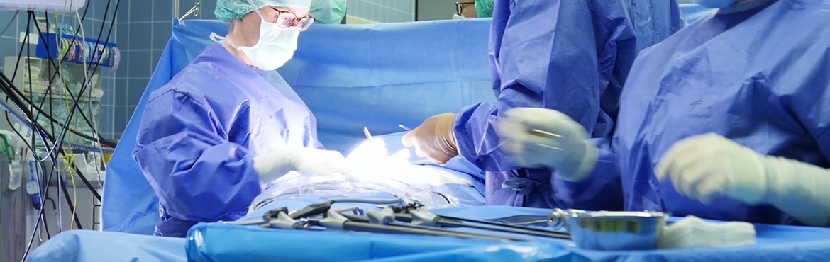 Blick in einen Operationssaal. Zu sehen ist ein auf dem Rücken liegender Patient, der mit einem blauen Tuch abgedeckt ist. Davor steht ein Tisch der mit einem blauen Tuch abgedeckt ist und auf dem operationsbesteck liegt. Um den Patienten stehen fünf Ärzte und Schwestern die die Operation durchführen.