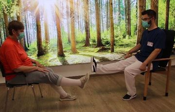 Der Pysiotherapeut Sören Schulz und eine Patienten sitzen sich auf Stühlen gegenüber und halten beide das rechte Bein gestreckt in die Luft. Hinter ihnen ist ein großes beleuchtetes Wandbild mit der Sicht aus einem Wald auf eine sonnendurchflutete Lichtung zu sehen.