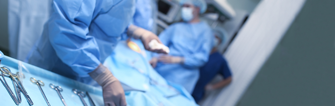 Eine Frau in einem sterilen Op-Kittel steht vor einem mit einem blauen Tuch abgedeckten Tisch mit Op-Besteck. Im Hintergrund sieht man verschwommen einen Patienten mit blauen Tüchern abgedeckt auf einem Op-Tisch und Ärzte die um ihn herum stehen und eine Operation durchführen.